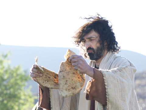 Depois de instruir a multidão a sentar-se no chão, Jesus pegou os sete pães e os peixes e, depois de dar graças, Ele os partiu em pedaços. – Slide número 7